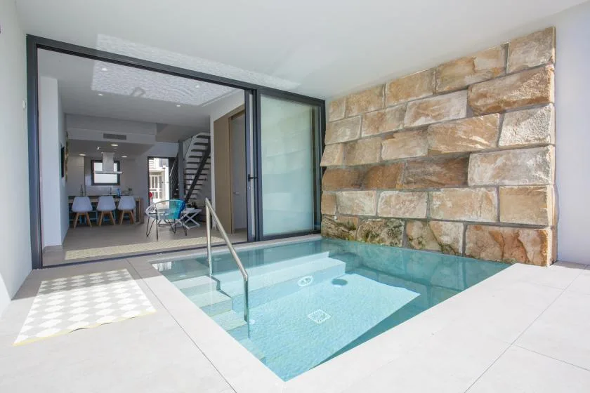 Una pequeña piscina interior junto a un muro de piedra y puertas de vidrio abiertas.