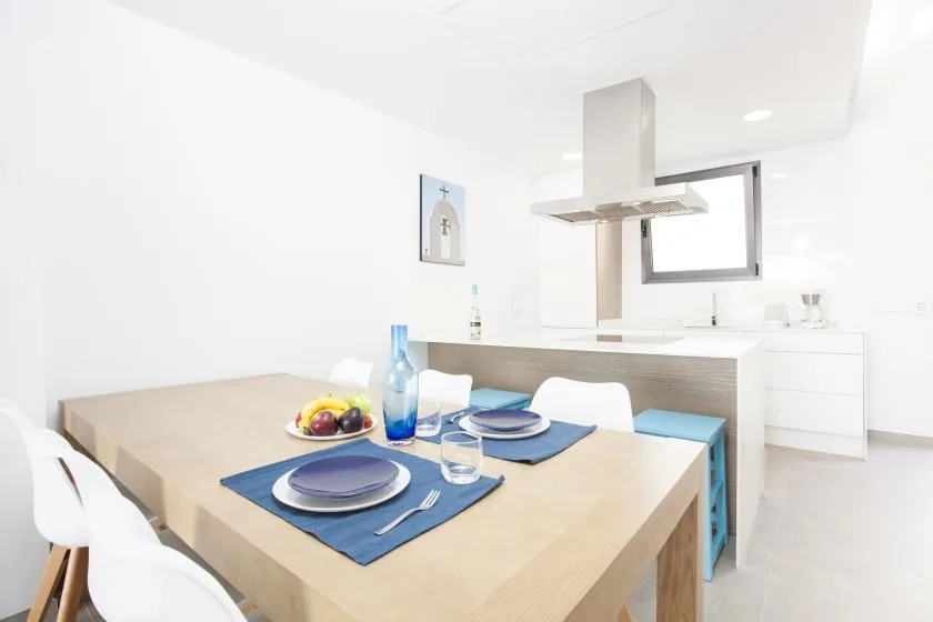 Una mesa de comedor con cubiertos está junto a una cocina abierta y luminosa.