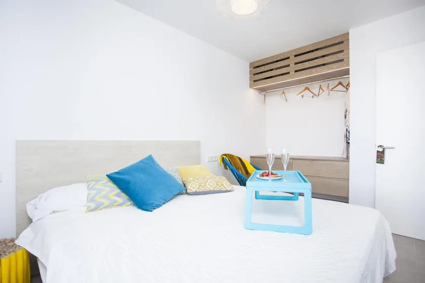 Un dormitorio con una cama blanca, cojines coloridos y una pequeña mesa azul.