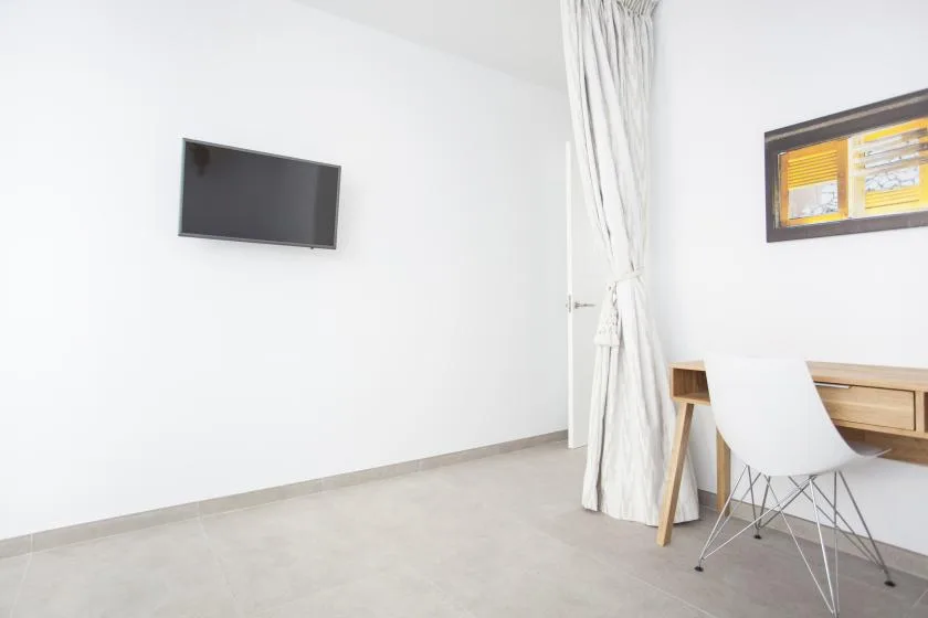 Una habitación luminosa con un televisor en la pared y un escritorio junto a una ventana con cortinas.