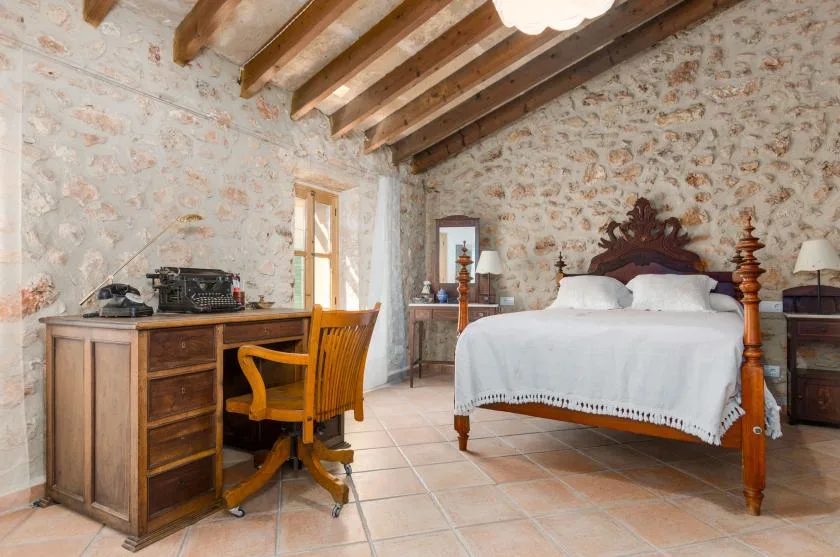 A rustic bedroom with a wooden bed and a desk beneath exposed ceiling beams.