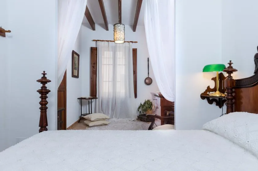 A bedroom with a white bed, sheer curtains, and exposed wooden ceiling beams.