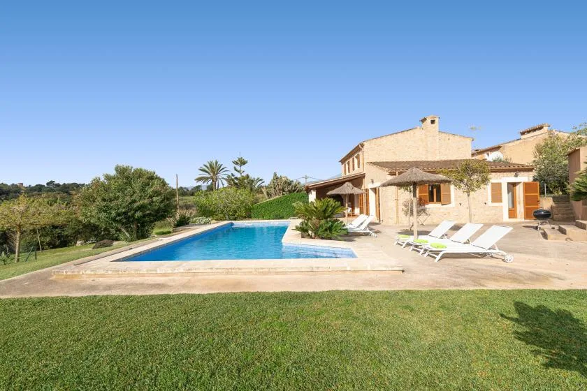 Una casa de piedra da a una piscina y un césped bajo un cielo despejado.