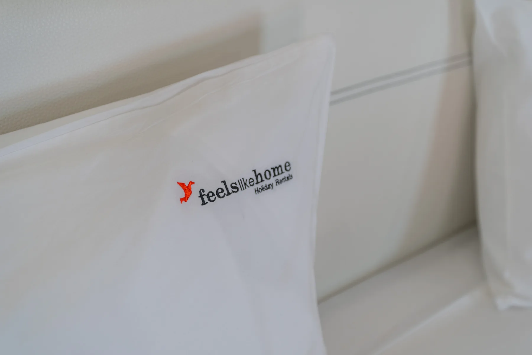 Una almohada blanca con un pequeño logotipo está apoyada contra una pared clara.