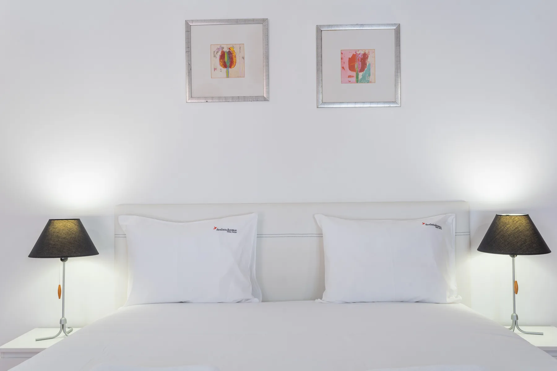 Un dormitorio con ropa de cama blanca, lámparas de noche y cuadros sobre la cama.
