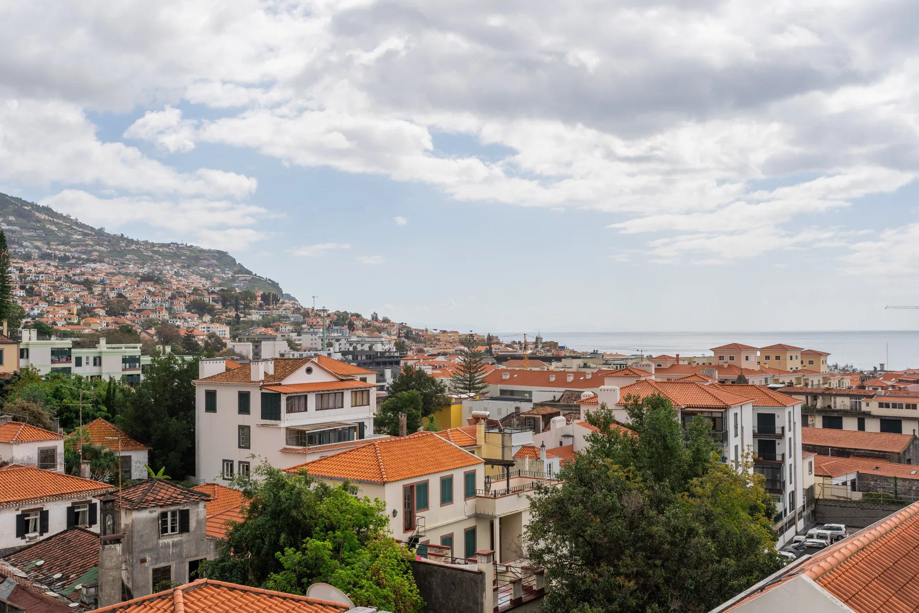 A coastal cityscape with red-roofed buildings and hills beneath a cloudy sky.