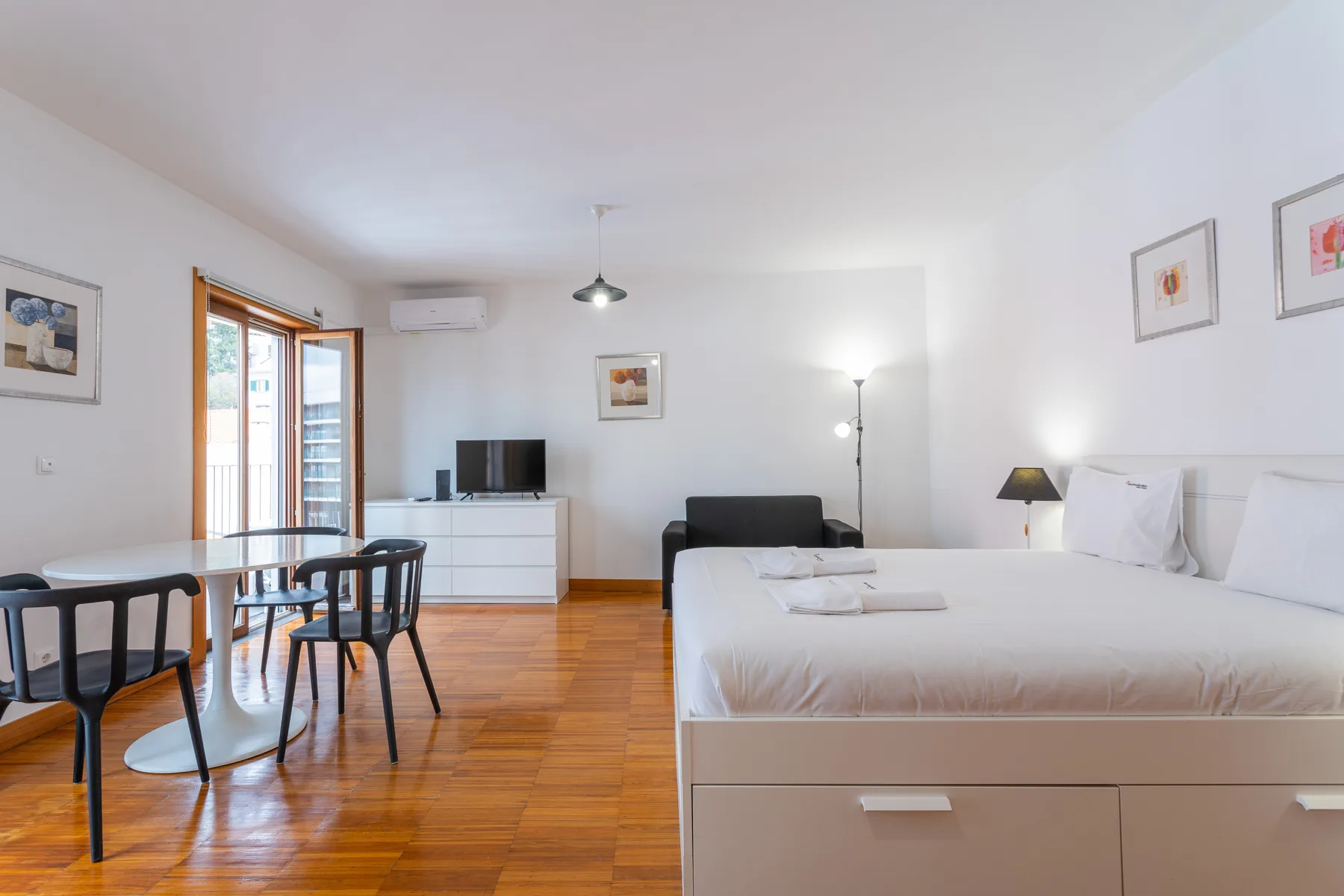 A bright studio bedroom with a bed, seating area, and dining table beside the balcony doors.