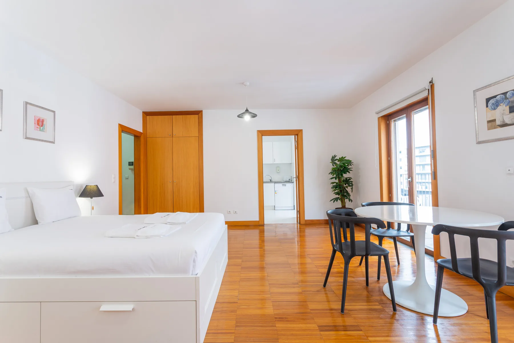 A bright studio room combines a bed, dining table, and wooden flooring.