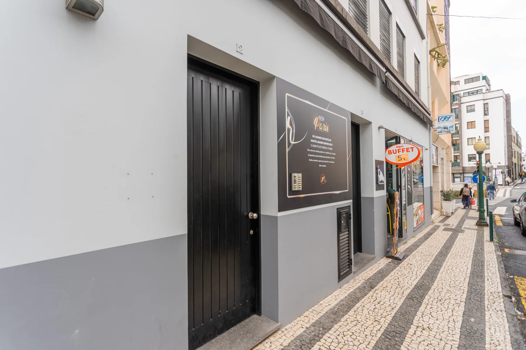 A city sidewalk runs past a building entrance and storefronts.