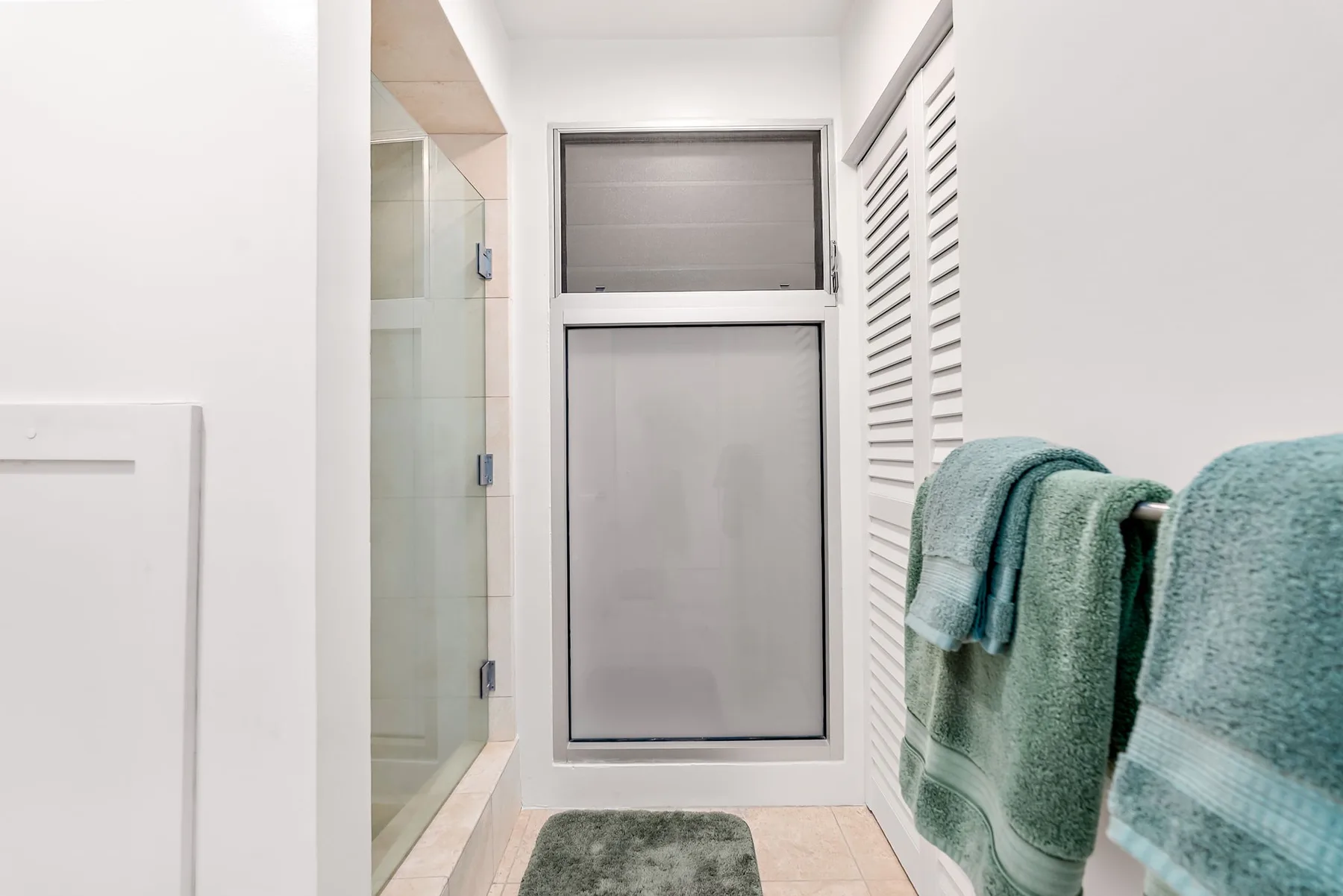 Un baño luminoso con ducha de vidrio, ventana translúcida y toallas colgadas.