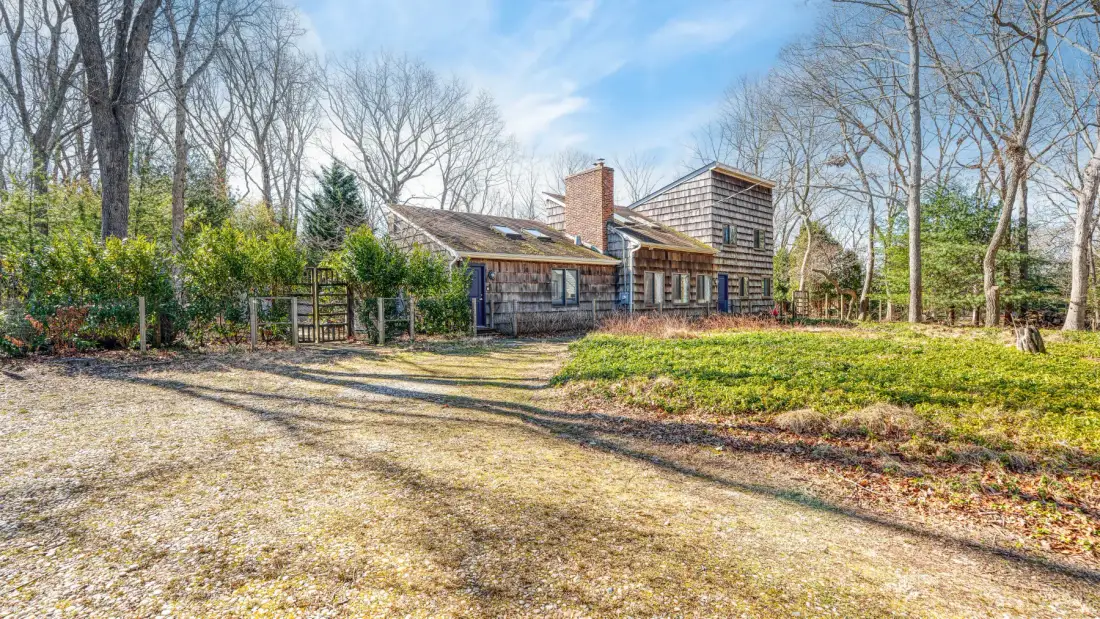 A house sits beyond a broad gravel driveway, surrounded by leafless trees.