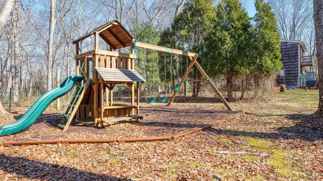 A wooden outdoor play structure with a slide stands in a leaf-covered yard.