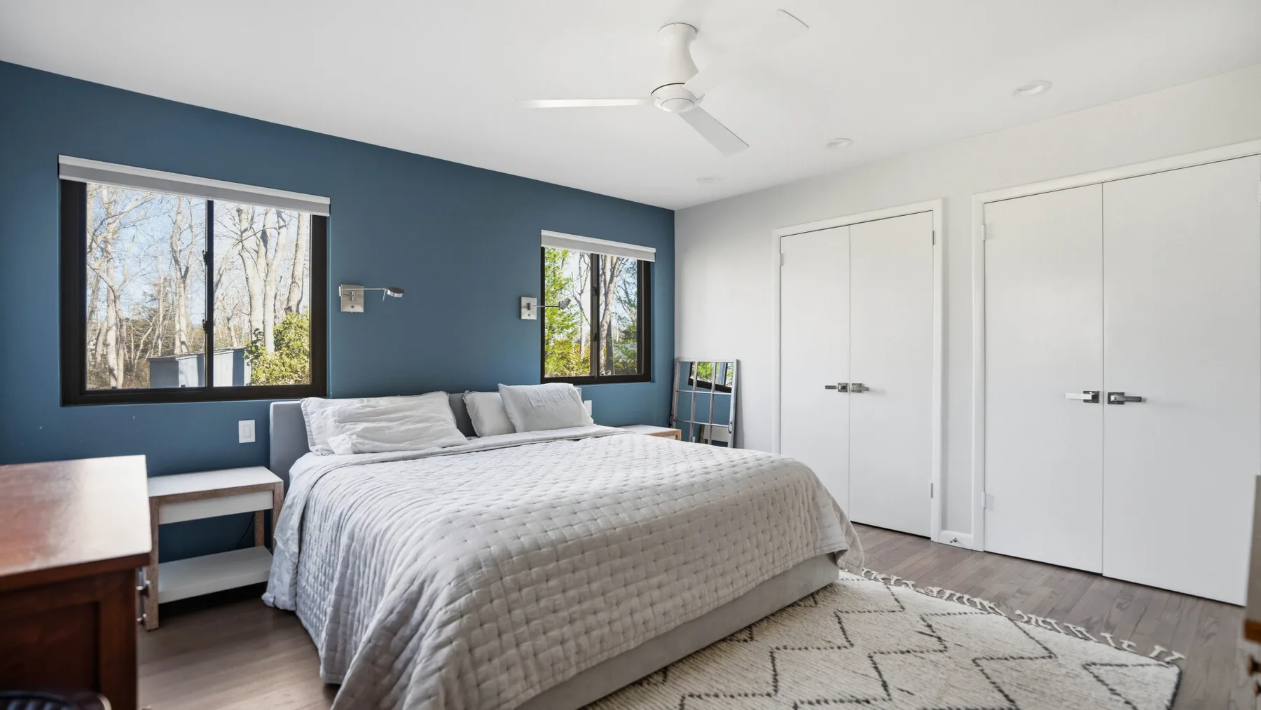 A bedroom with a large bed, blue accent wall, and two windows.