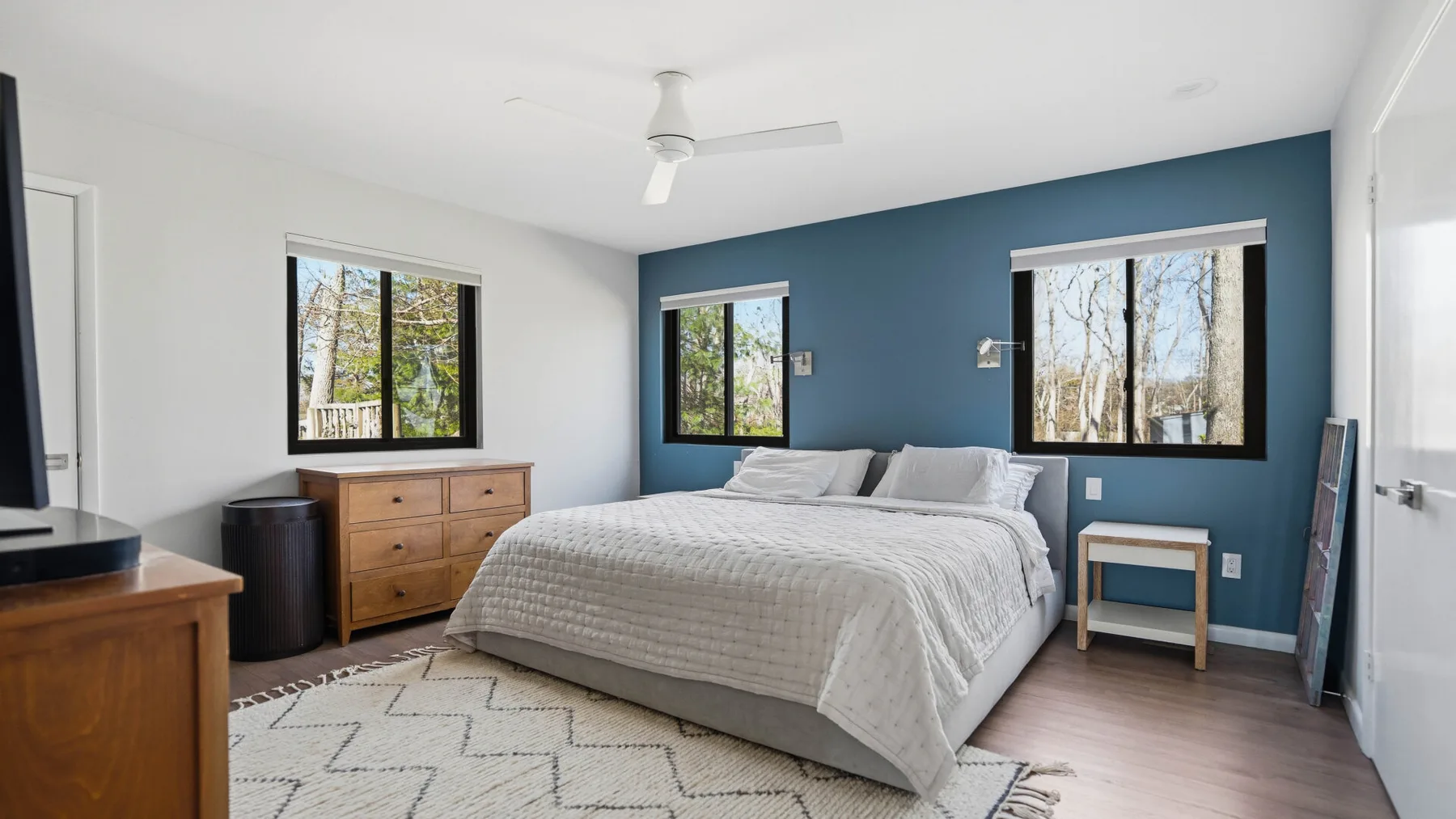 A bedroom with a bed against a blue accent wall and windows overlooking trees.