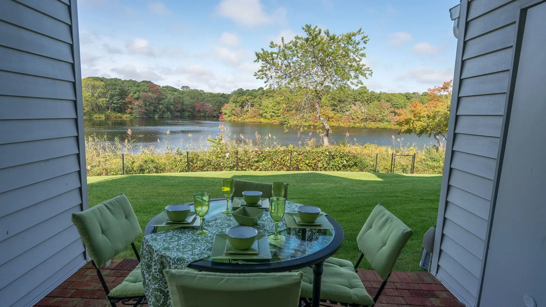Una mesa de comedor al aire libre en un patio da a un césped, un lago y árboles.