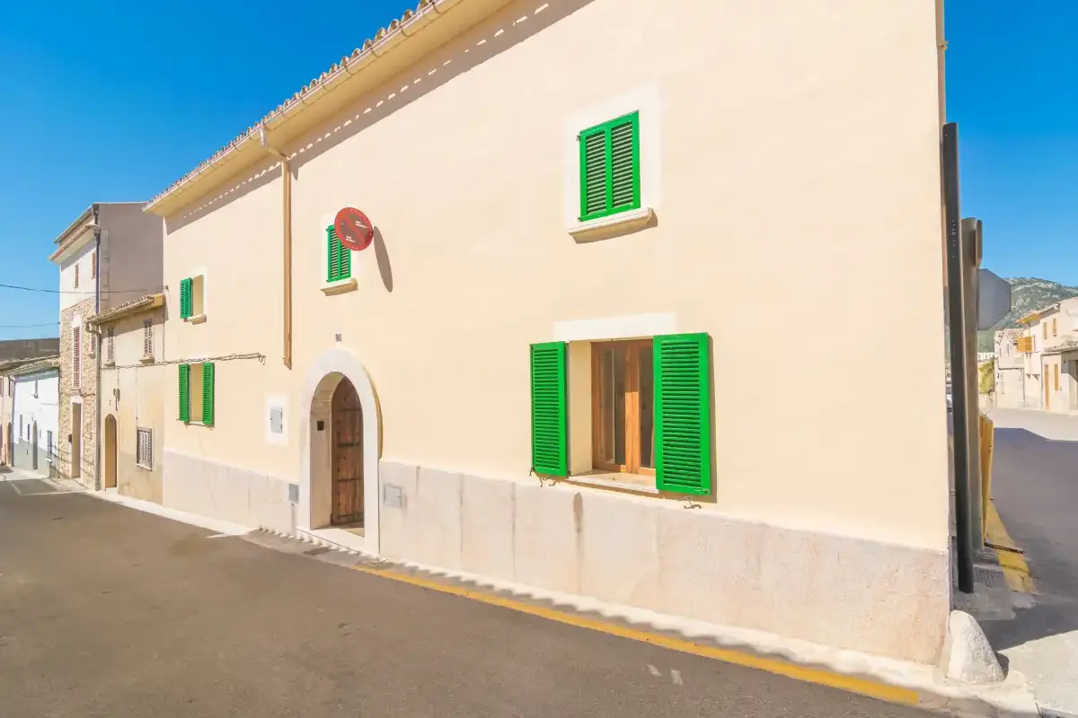 A sunlit building facade with green shutters and an arched doorway along a street.
