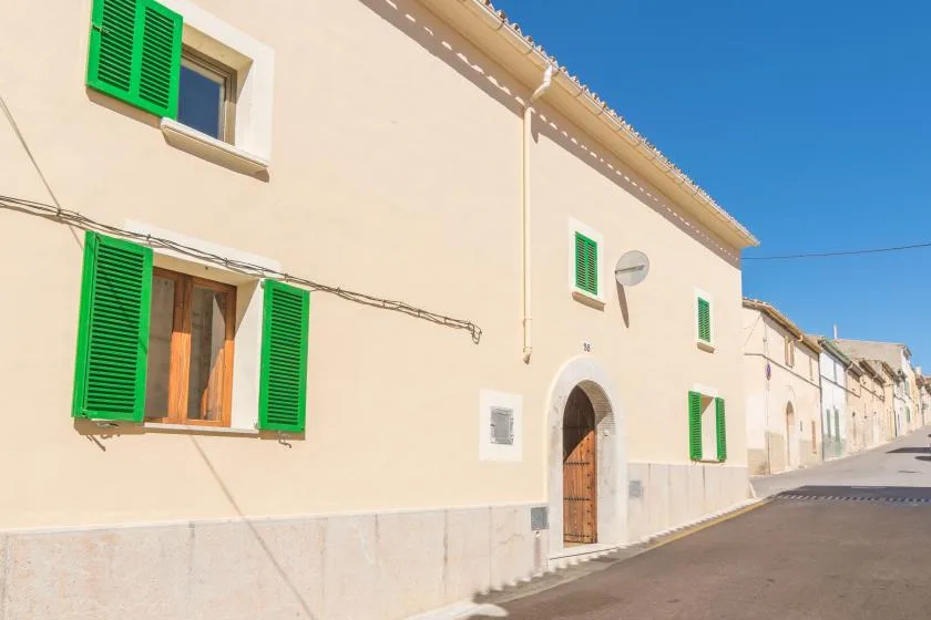 A sunlit street of pale buildings with green shutters and an arched doorway.