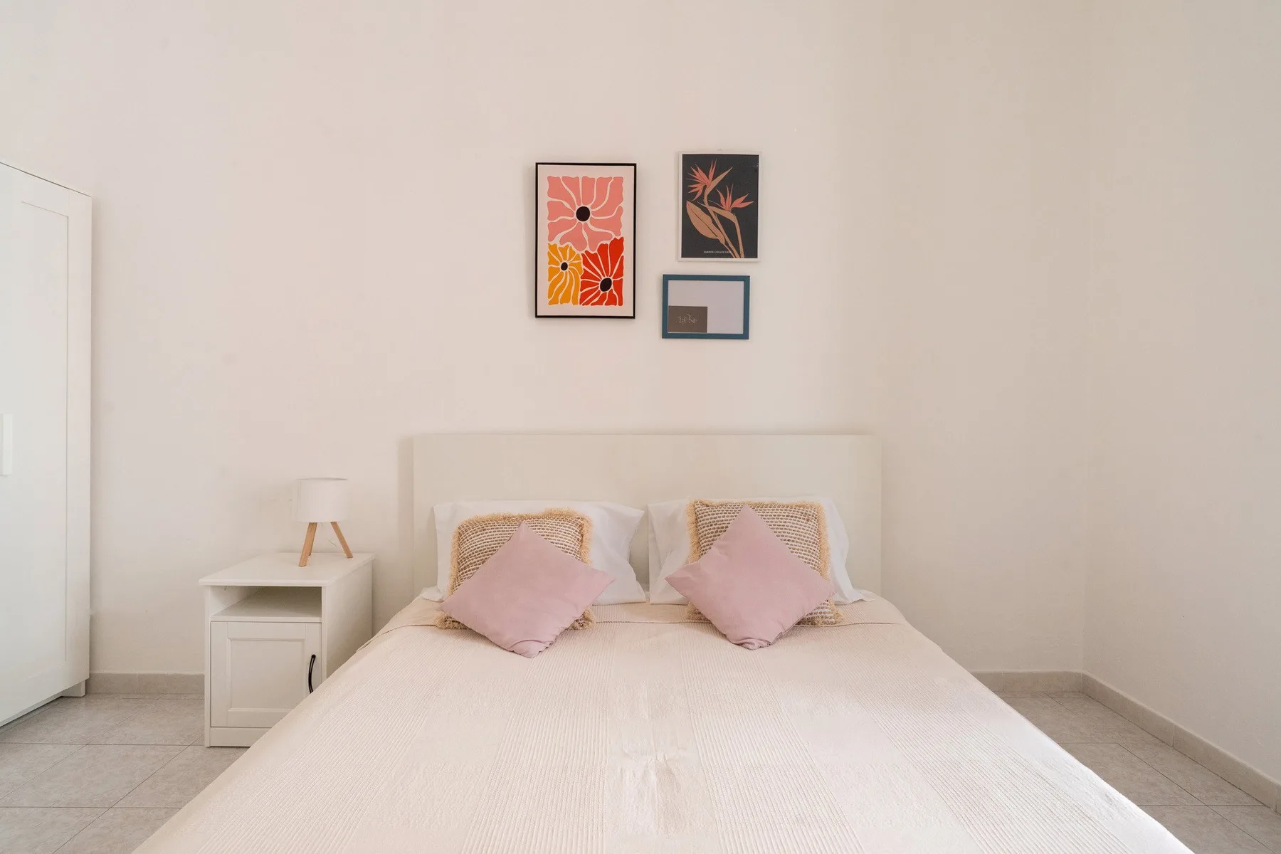 A bright bedroom features a bed with pink pillows and framed floral artwork.