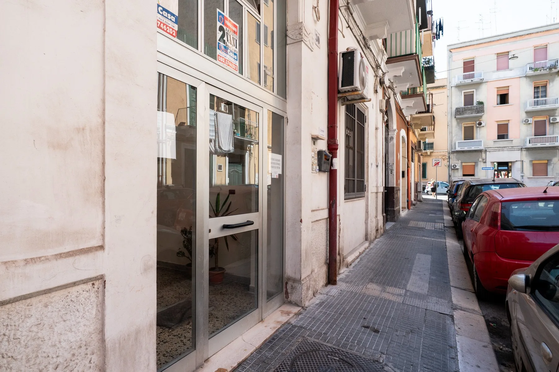 A narrow urban sidewalk runs between building entrances and parked cars.