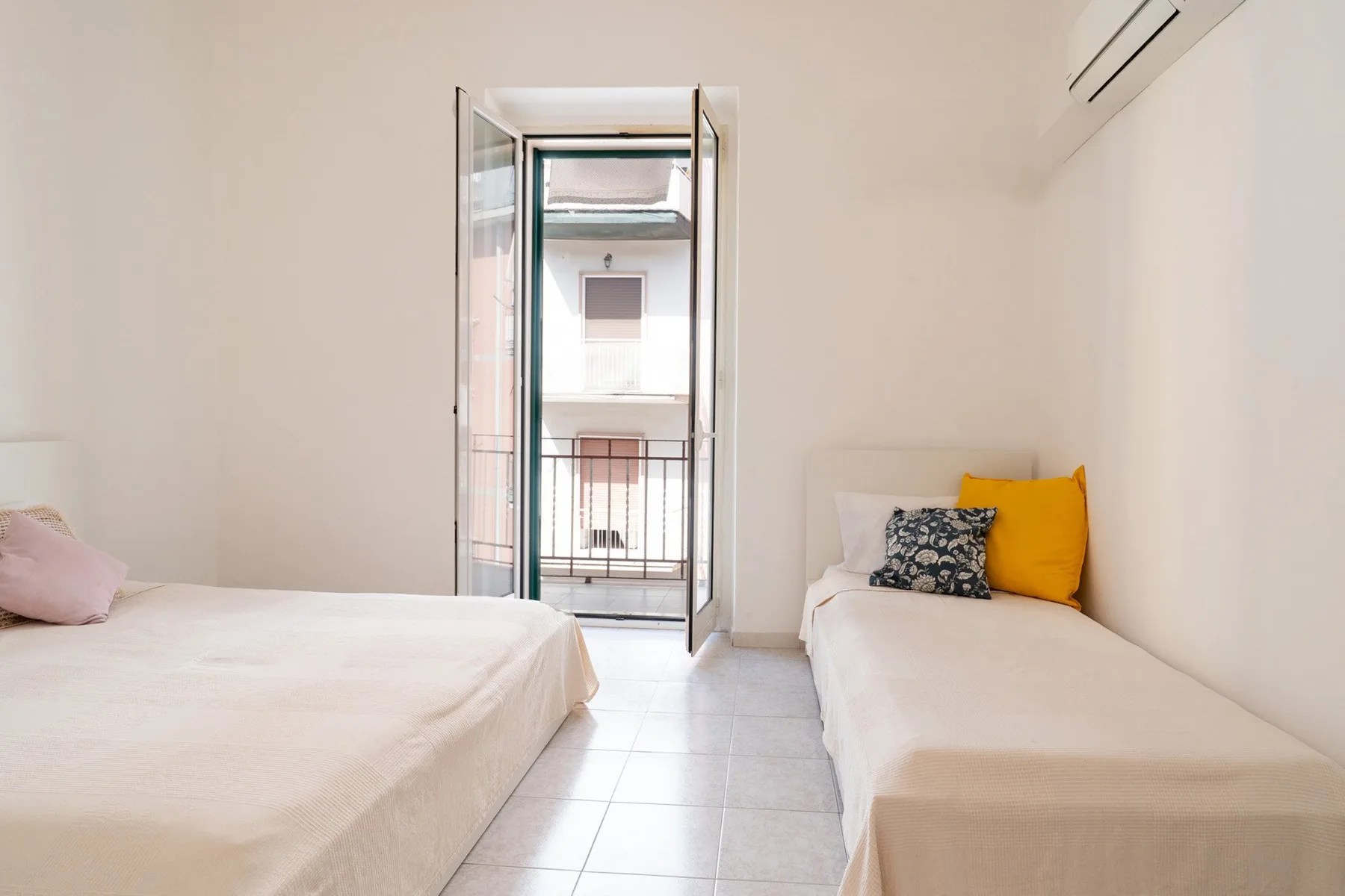 A bright bedroom with two beds and an open balcony doorway.