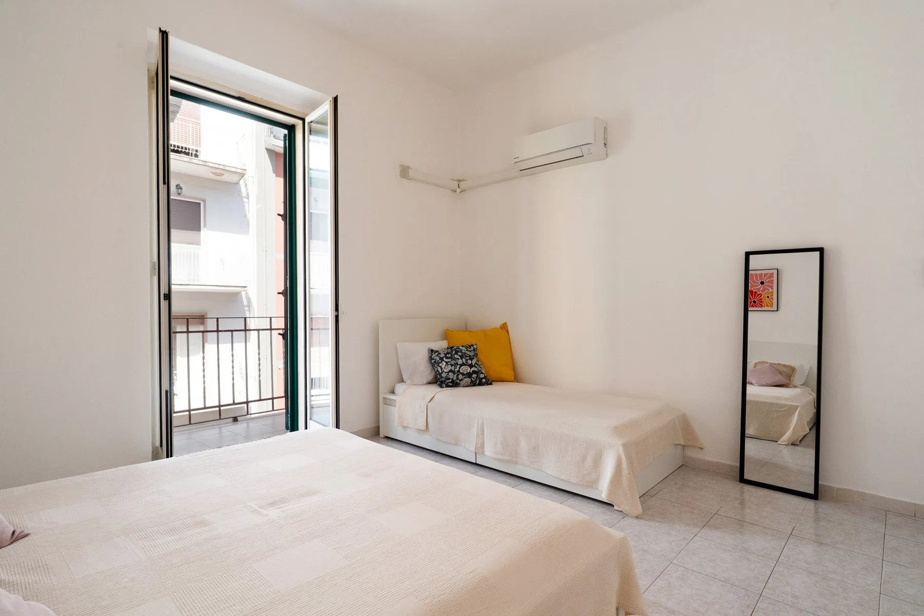 A bright bedroom with two beds, a tall mirror, and an open balcony door.