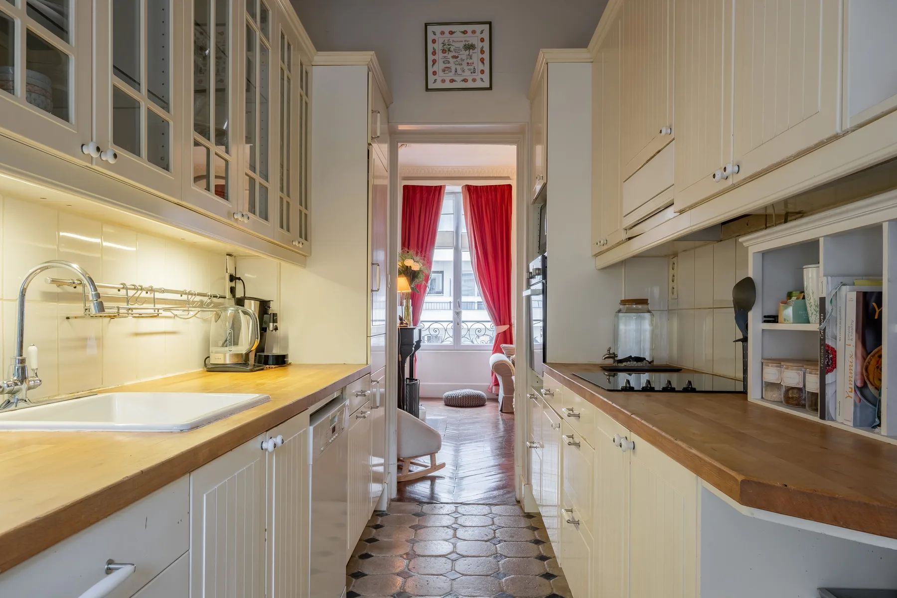 Una cocina estrecha con armarios y encimeras da a una habitación luminosa con cortinas rojas.