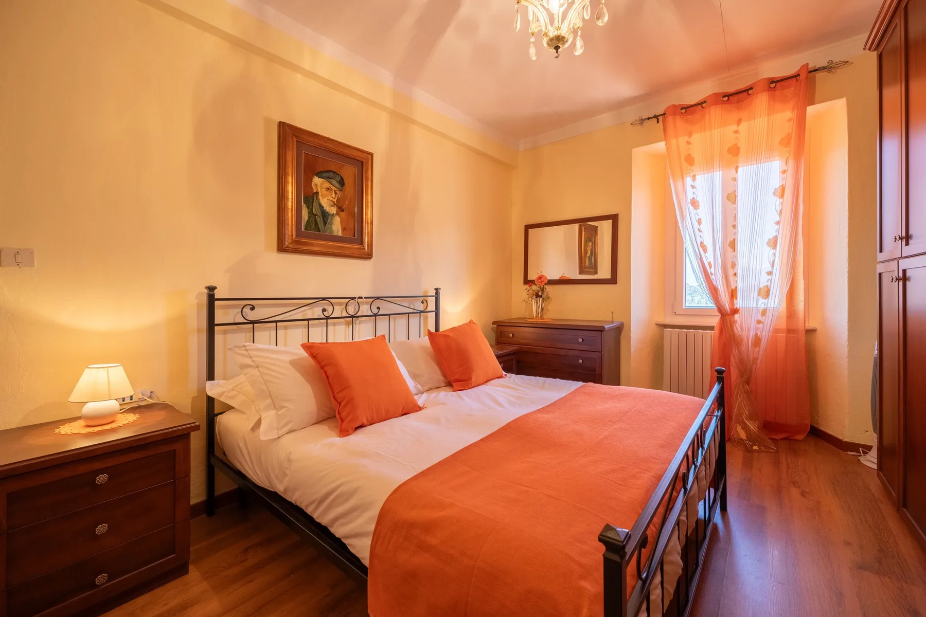 A bedroom with a metal bed, orange bedding, and a sunlit window.