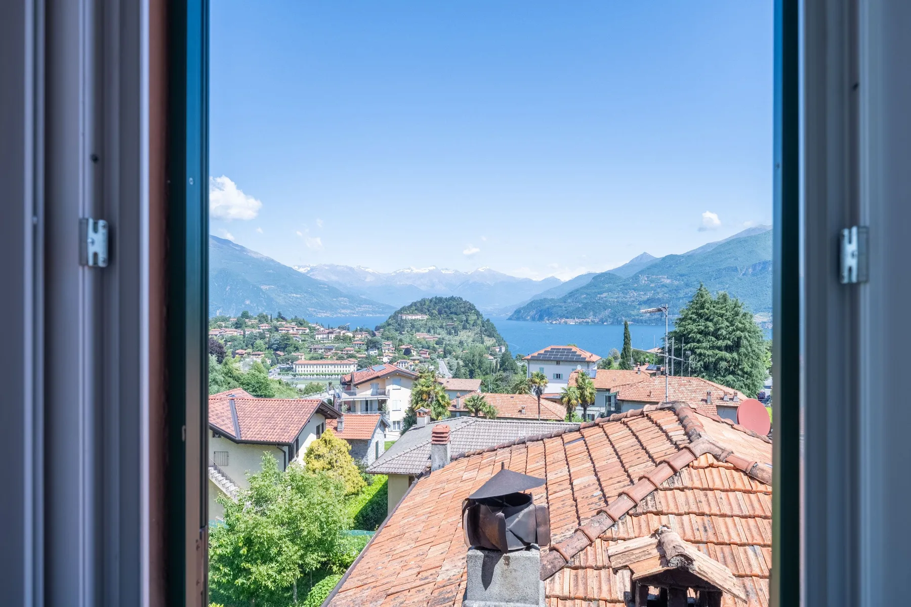 An open window overlooks rooftops, a lakeside town, and surrounding mountains.