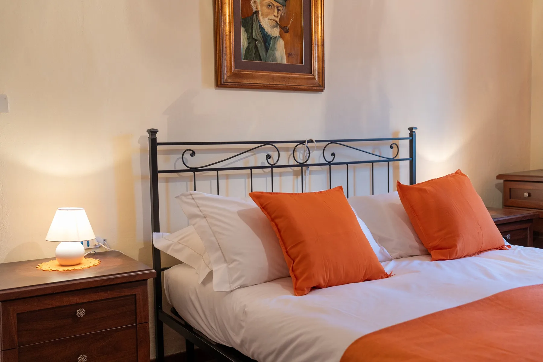A bedroom with a neatly made bed, orange pillows, and a framed portrait above the headboard.
