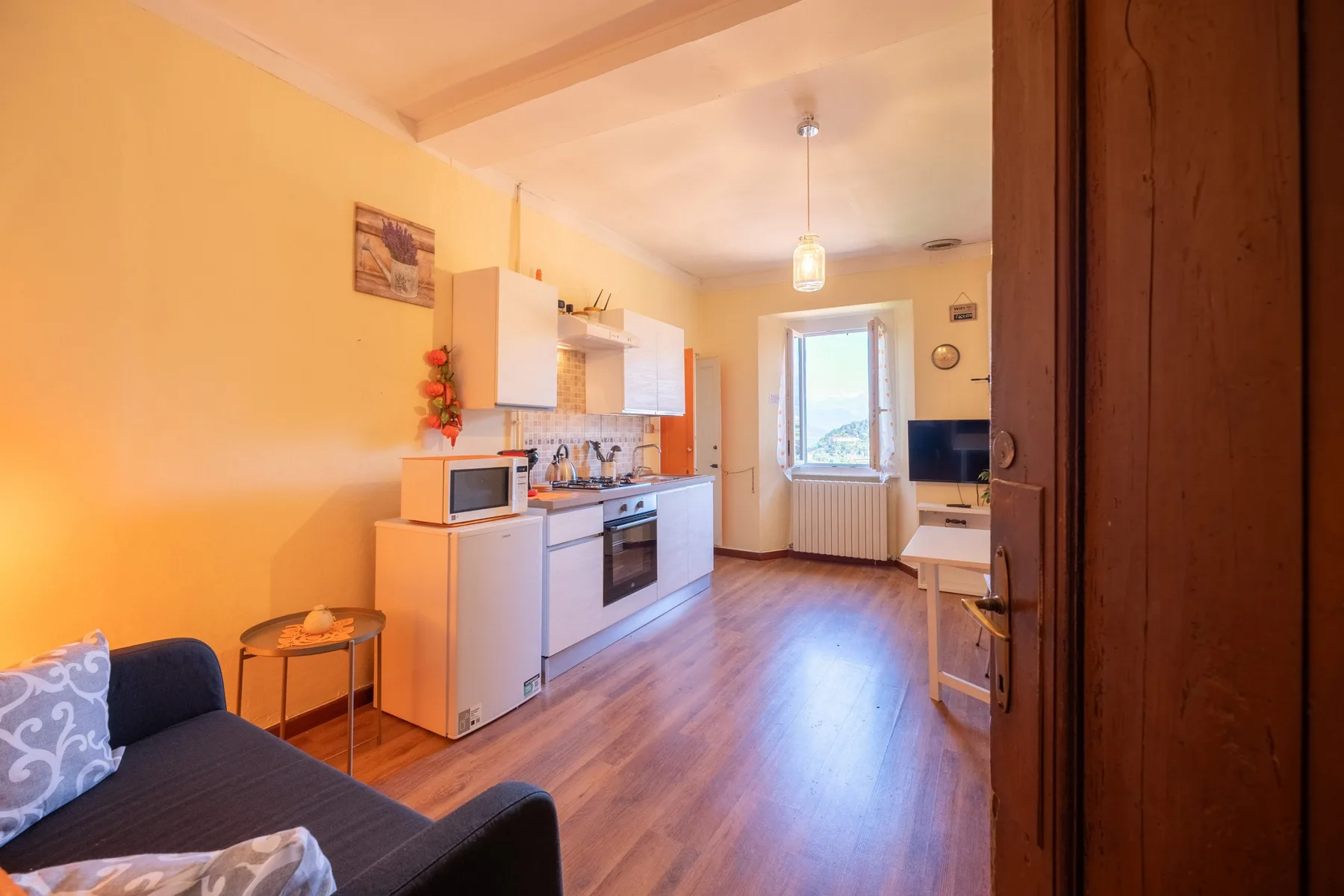 A bright kitchen and living area with wooden flooring and a window at the far end.