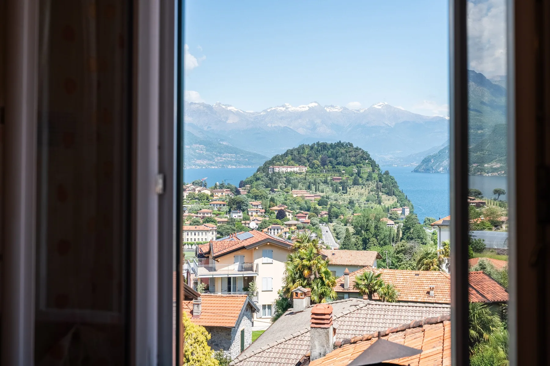 A view through an open window overlooks hillside homes, a lake, and distant mountains.