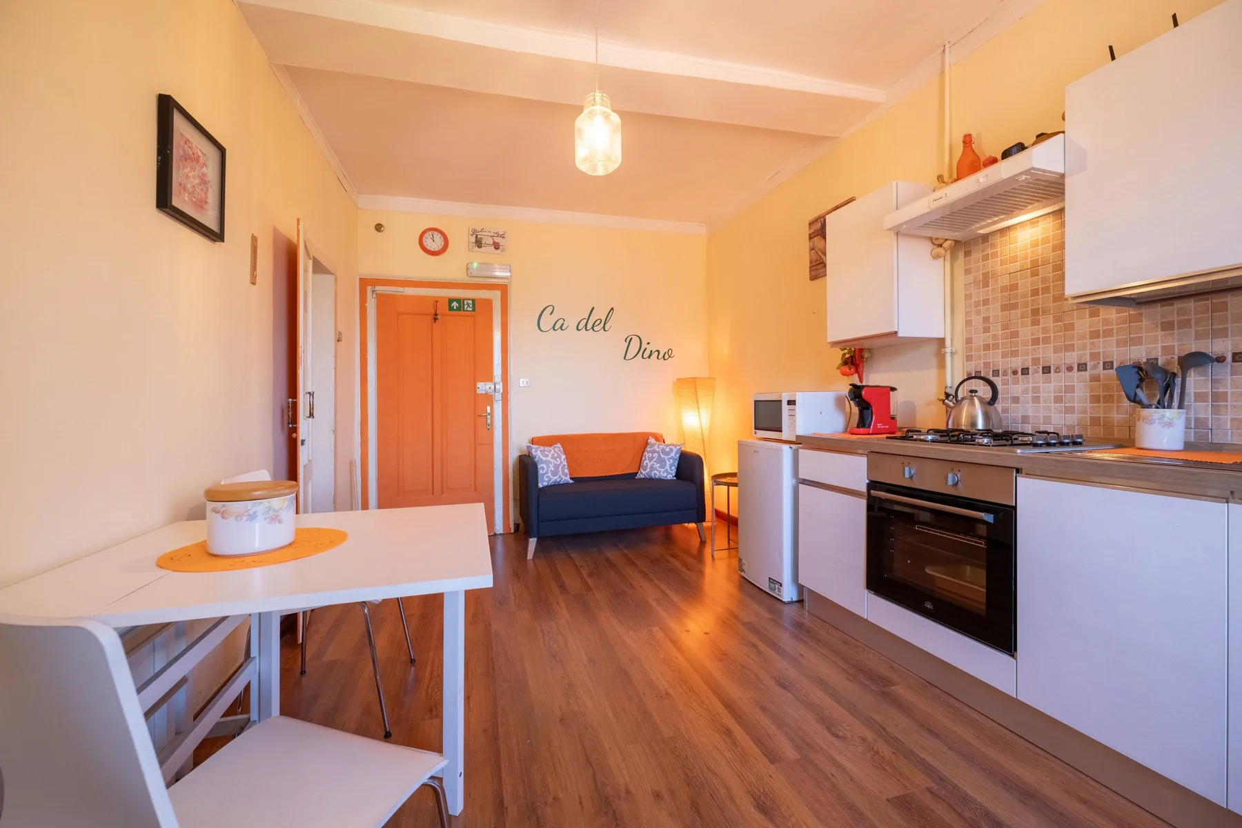 A bright studio room combines a small seating area, dining table, and kitchenette.