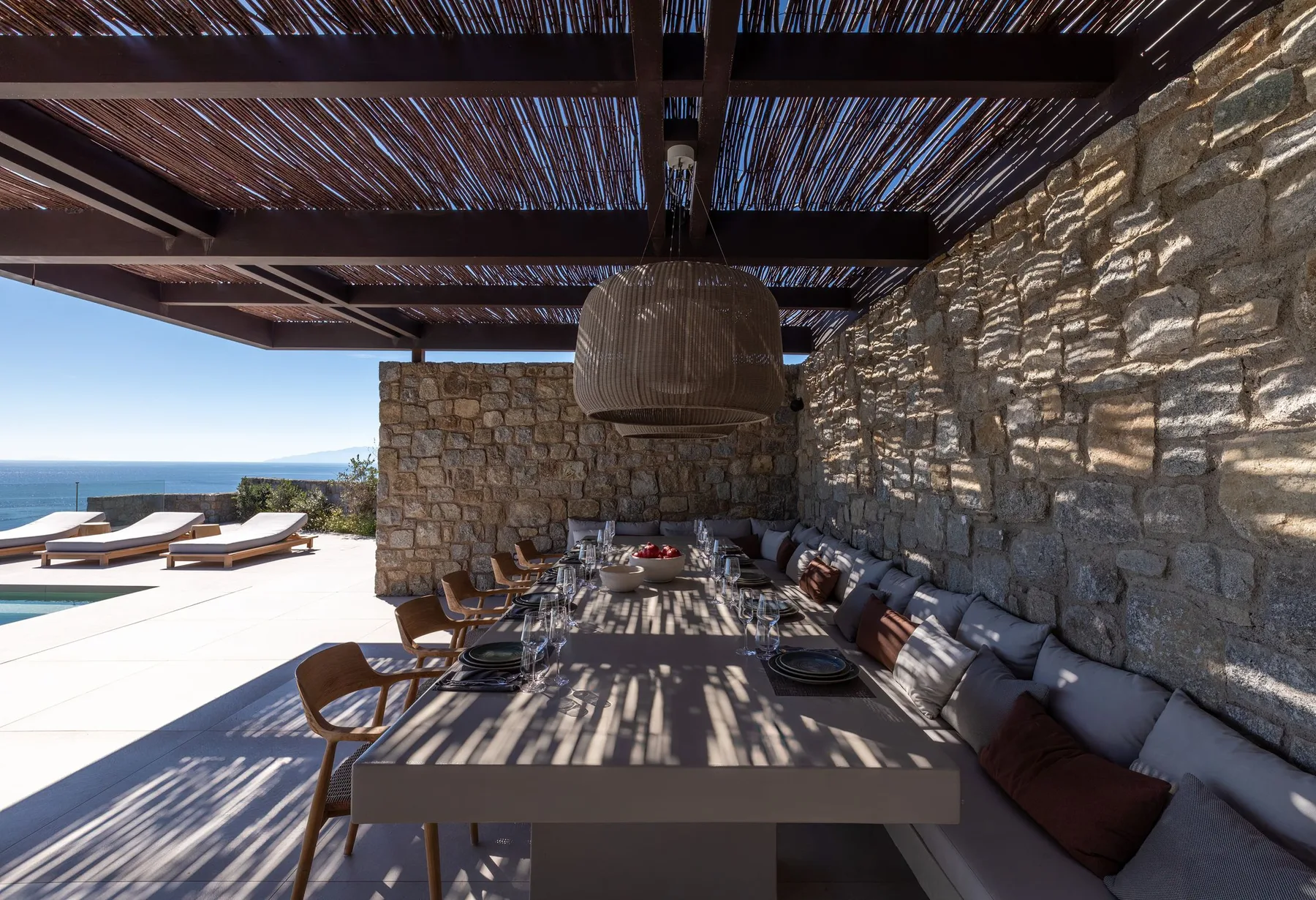 A shaded outdoor dining area with seating beside a stone wall and a pool.