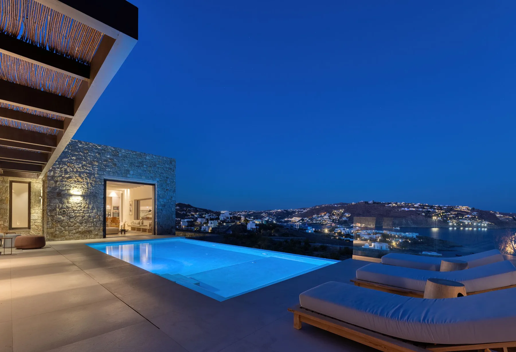 An outdoor pool and lounge seating overlook a coastal town at dusk.