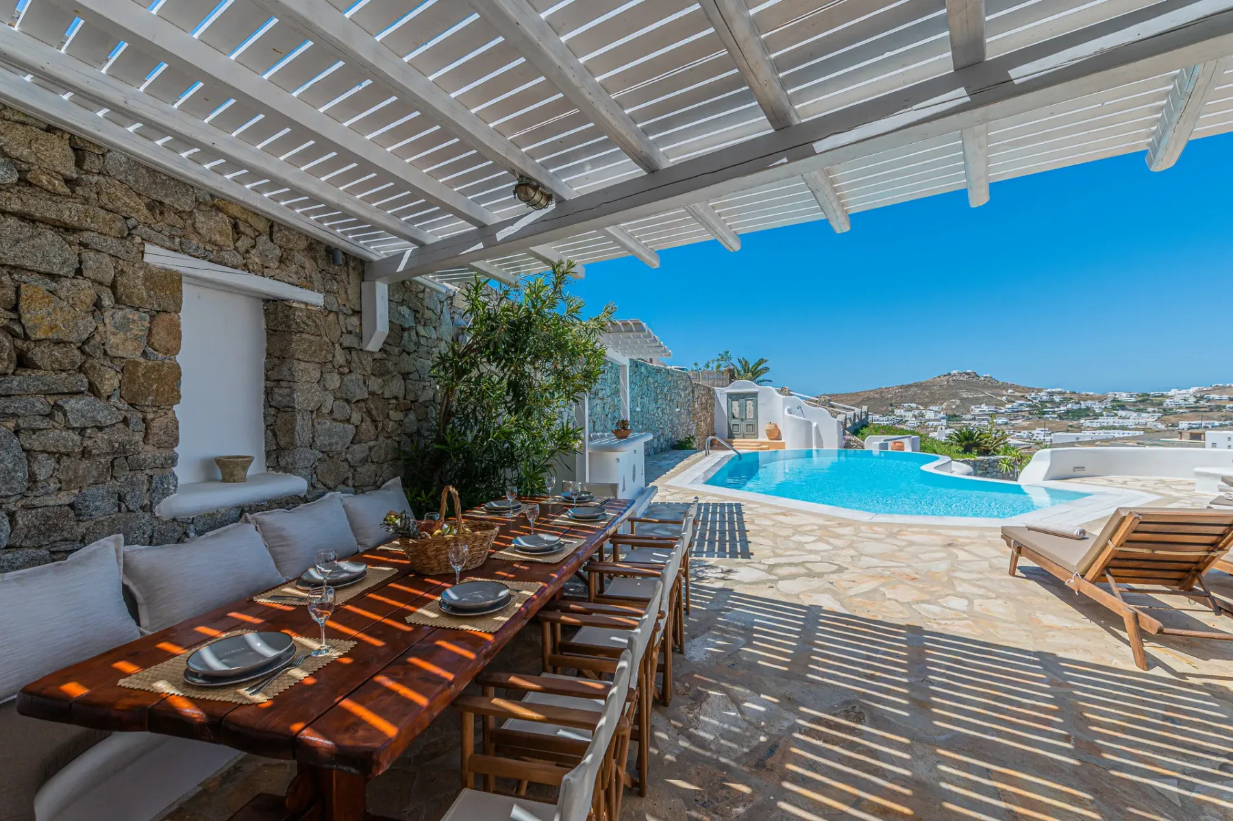 A shaded outdoor dining area beside a swimming pool, with a hillside view.