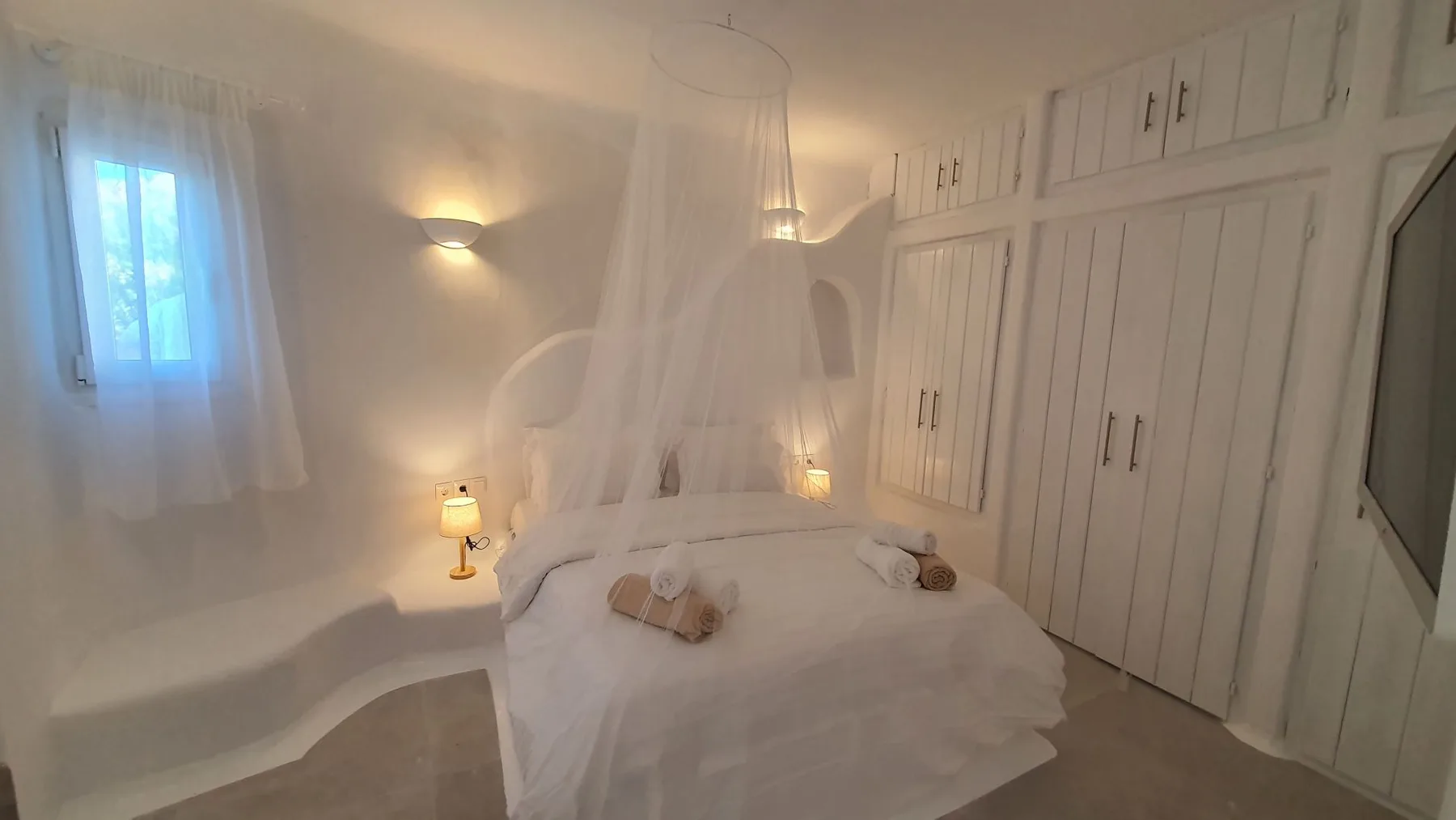 A bedroom with a bed draped in white fabric and built-in wardrobes.