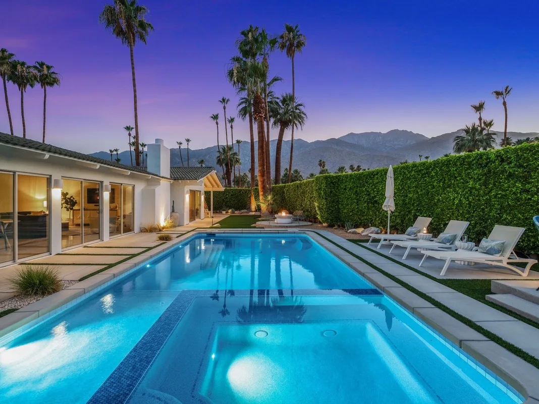 Una piscina en el patio junto a una casa moderna, con asientos reclinables y palmeras.
