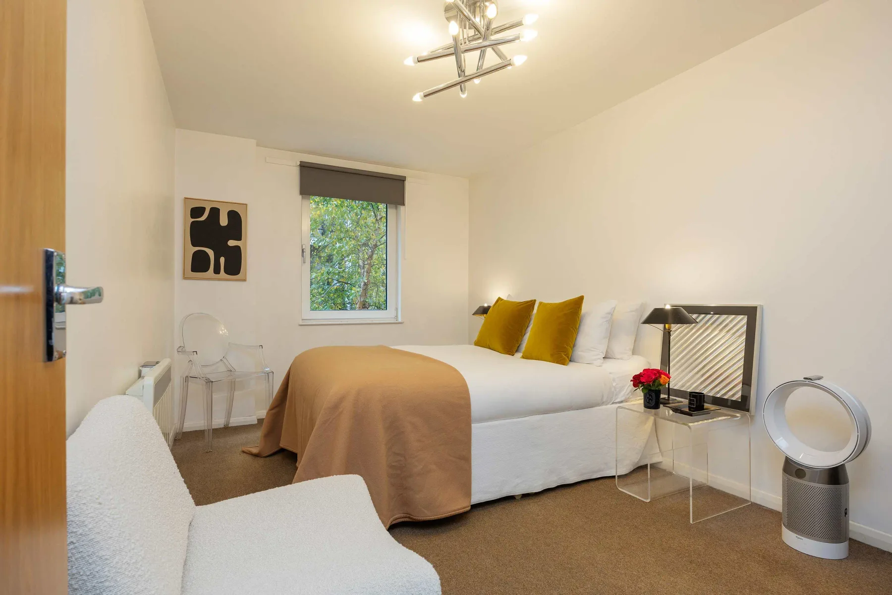 A bedroom with a bed, white seating, and a window overlooking greenery.