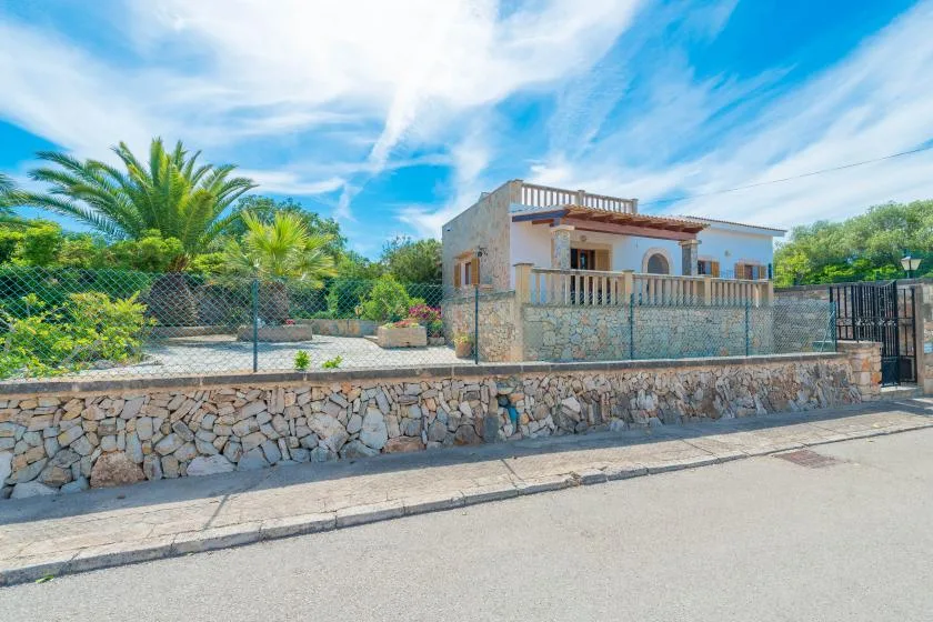 Una casa de color claro con balcón se encuentra detrás de un muro de piedra y un jardín.