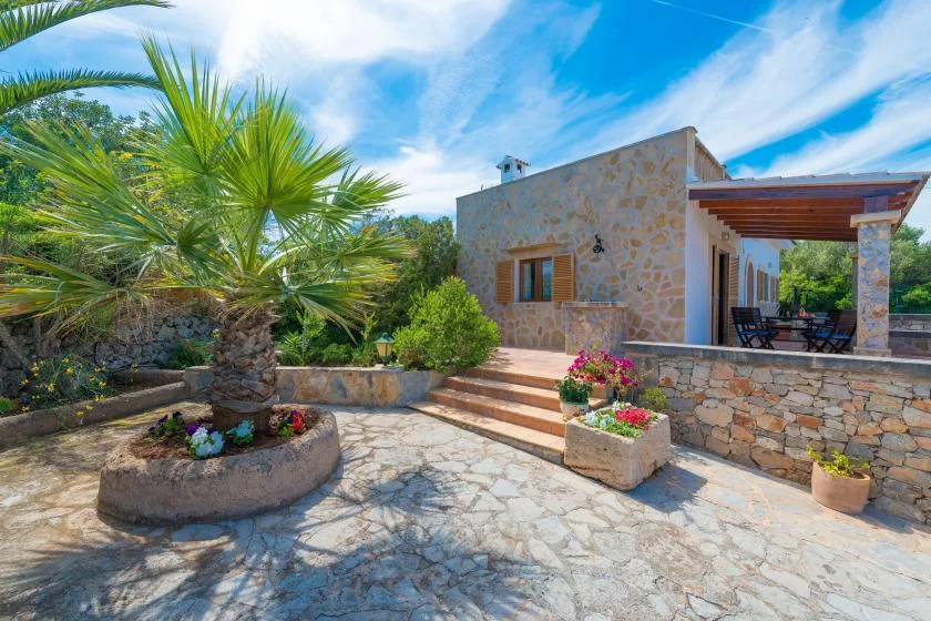 Una casa de piedra da a un patio pavimentado con jardineras y una terraza sombreada.