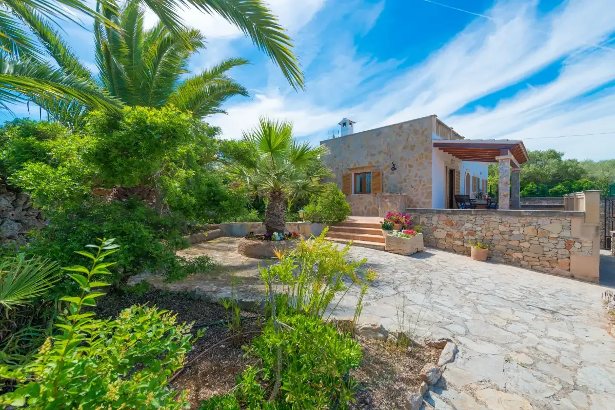Una casa de piedra y un patio pavimentado están rodeados de palmeras y plantas.