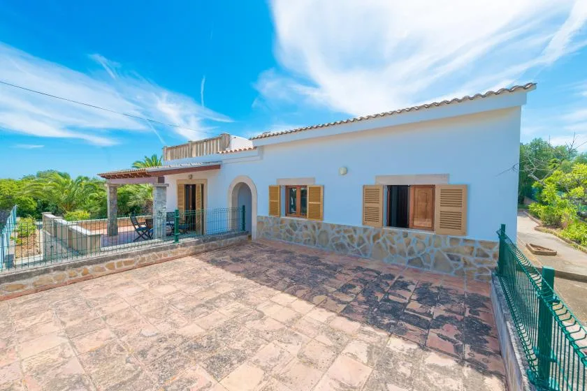 Una terraza exterior soleada junto a una casa blanca con ventanas de contraventanas.