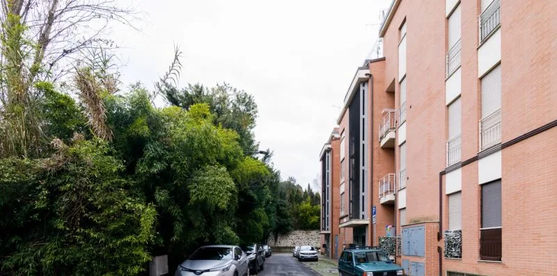 A street runs beside a row of brick apartment buildings and leafy trees.
