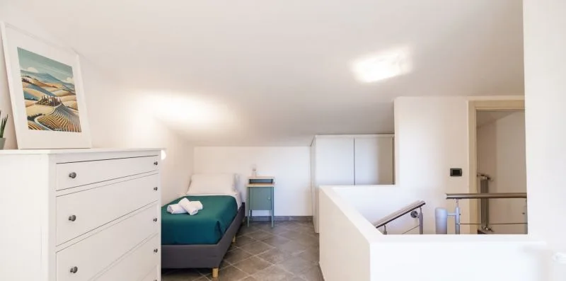 A bright loft bedroom has a bed beside a white dresser and stair railing.