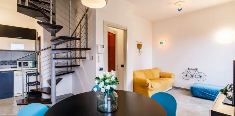An open-plan living area with a yellow sofa, dining table, and spiral staircase.