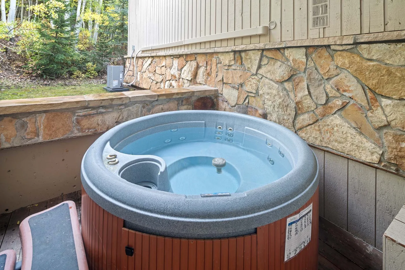 An outdoor hot tub sits beside a stone retaining wall and wooded area.