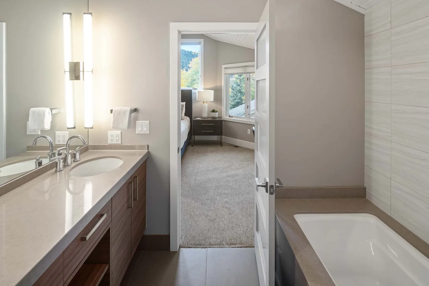 A bathroom vanity and bathtub overlook an open doorway to a carpeted bedroom.