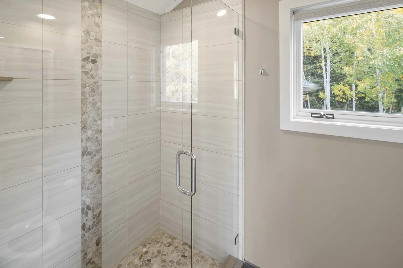 A tiled bathroom shower sits beside a window overlooking leafy trees.