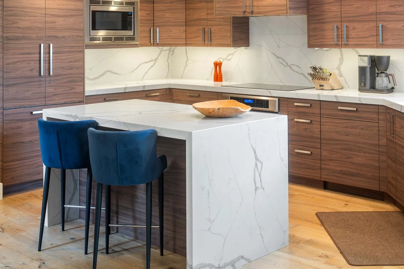 A modern kitchen with wood cabinetry, a marble island, and blue seating.