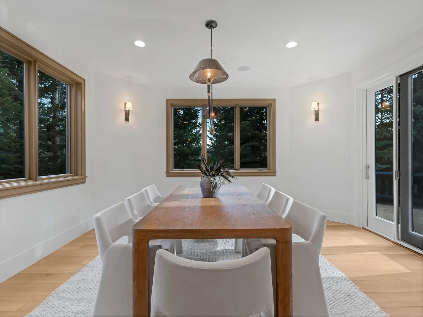 A bright dining room with a wooden table, white seating, and large windows.
